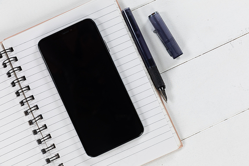 Close up top view of a black smartphone, a notebook and a black pen arranged on a plain white background