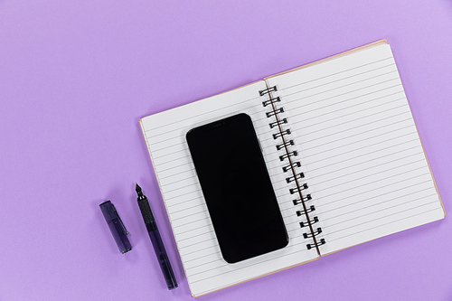 Close up top view of a black smartphone, a notebook and a black pen arranged on a plain purple background