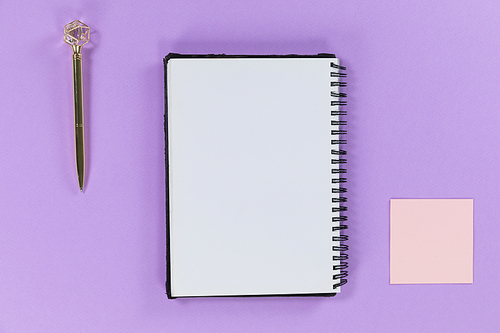 Close up top view of a notebook a pen and a small pink sheet of paper arranged on a plain purple background