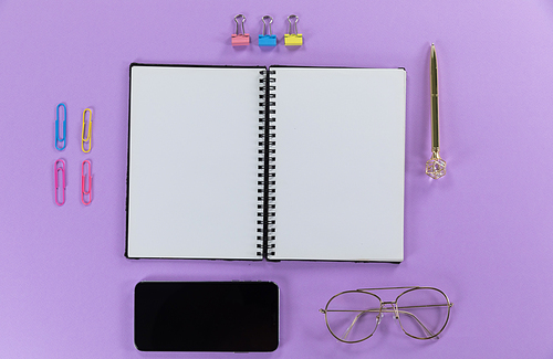 Top view of a black smartphone, a notebook, a pen, glasses and colorful paperclips arranged on a plain purple background