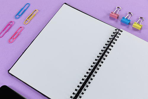 Close up top view of an empty page in a notebook and colorful paperclips arranged on a plain purple background