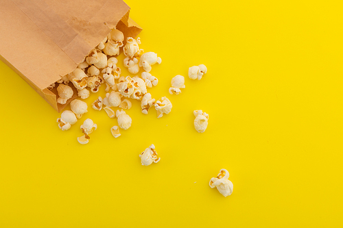 High angle view of popcorn spilling out of brown paper bag on yellow background. fun party food sweet treat snack copy space concept.