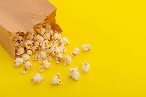 High angle view of popcorn spilling out of brown paper bag on yellow background. fun party food sweet treat snack copy space concept.
