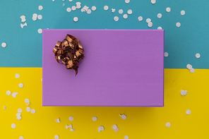 Purple gift box with ribbon and white confetti on yellow and blue background. happy birthday party celebration giving receiving concept.