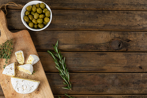 Top view of a wooden cutting board with cheese arranged on a on a textured wooden surface with a spring of thyme and a bowl of green olives.