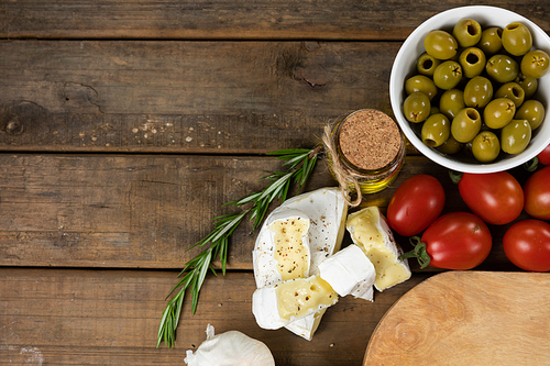 Top view of a wooden cutting board with a bowl of green olives, some cheese, cherry tomatoes and a spring of fresh thyme, arranged on a on a textured wooden surface.