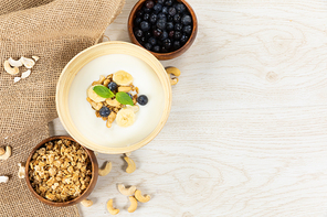 Top view of three bowls with muesli, blueberries and yoghurt with fruits and nuts, arranged on a textured white wooden surface.