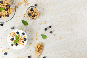 Top view of three bowls with muesli, nuts, blueberries and yoghurt, arranged on a textured white wooden surface.