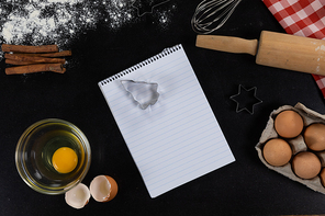 Top view of an empty notebook page with ingredients prepared for baking cookies, arranged on a plain black surface.