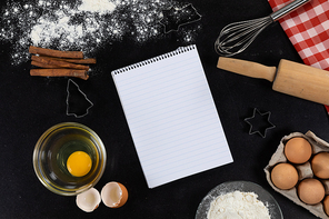 Top view of an empty notebook page with ingredients prepared for baking cookies, arranged on a plain black surface.
