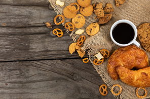 Top view of some cookies, pretzels and croissants put on a table cloth with a cup of black coffee, arranged on on a textured wooden surface.