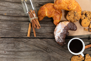 Top view of some cookies and croissants put on a table cloth with a cup of black coffee and a bottle with cinnamon, arranged on on a textured wooden surface.