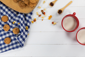 Top view of some cookies, spoons with dried fruits and nuts and two mugs with milk, arranged on on a textured white wooden surface with checkered tablecloth.