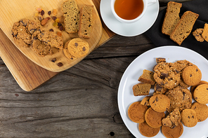 Top view of some cookies put on a white plate and a wooden cutting board, arranged on on a textured wooden surface with a cup of tea.