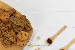 Top view of some cookies put on a wooden cutting board, arranged on on a textured wooden surface with two spoons with dried fruits.