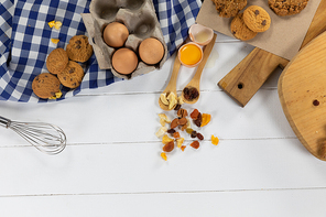 Top view of some cookies, spoons with dried fruits and eggs, arranged on on a textured white wooden surface with checkered tablecloth.