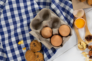 Top view of some cookies, spoons with dried fruits and eggs, arranged on on a textured white wooden surface with checkered tablecloth.