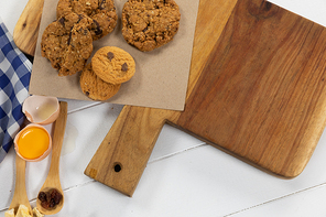 Top view of some cookies and ingredients prepared for baking, put on a wooden cutting board, arranged on on a textured white wooden surface with checkered tablecloth.