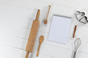 Top view of a composition with an empty page in notebook with cookie cutters, whisker and rolling pin, arranged on on a white textured wooden surface