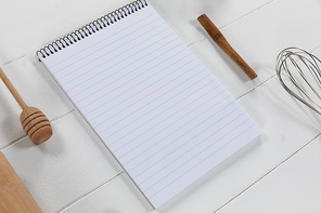 Top view of a composition with an empty page in notebook with wooden spoon and whisker, arranged on on a white textured wooden surface