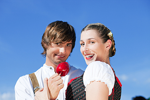 Young and beautiful couple in traditional Bavarian Tracht - Dirndl and Lederhosen - embracing each other on a fair like a Dult or the Oktoberfest eating traditional sugar apple