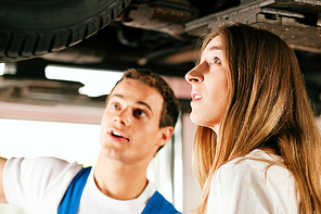 Woman talking to a car mechanic in his repair shop|both are standing underneath the auto which is lifted on a car hoist