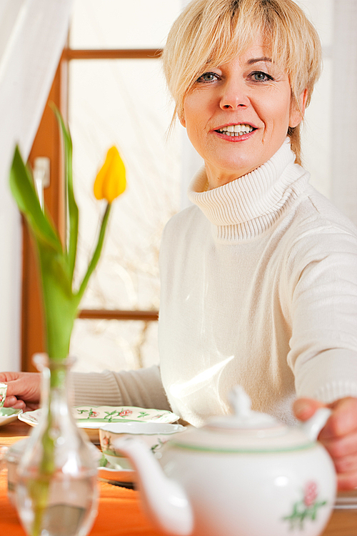 Woman sitting on a table for coffee or tea time grabbing for the coffee or tea pot|there are yellow tulip flower on the table|in the background is a window|whole scene is sunlit