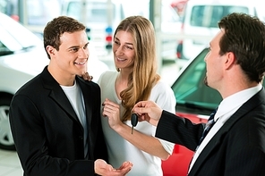 Sales situation in a car dealership|the dealer is handing auto keys to a young couple|they are excited|cars standing in the background