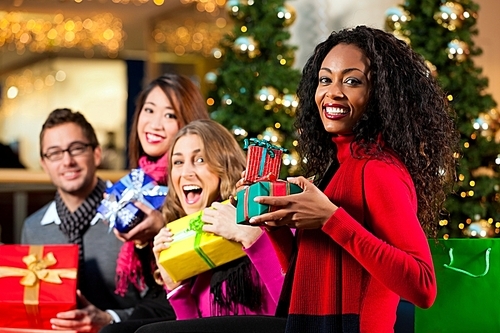 Diversity group of four people - Caucasian|black and Asian - sitting with Christmas presents and bags in a shopping mall in front of a Christmas tree with baubles