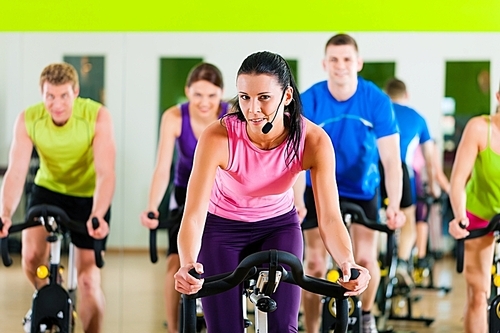 Group of five people - men and women - spinning in gym or fitness club exercising their legs doing cardio training; the trainer is in front