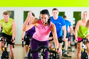 Group of five people - men and women - spinning in gym or fitness club exercising their legs doing cardio training; the trainer is in front