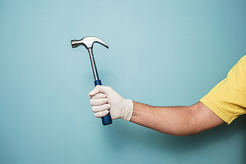 A young man’s hand wearing a latex glove is holding a hammer against a blue wall
