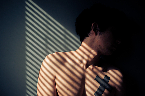 Sad naked man sitting by the window with shadows from blinds on his face