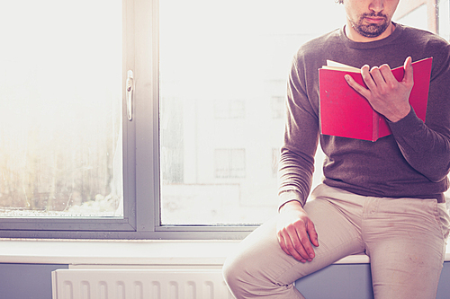 Young man sitting on window sill and reading a red book