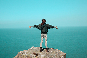 Young woman is standing on a cliff by the sea and raising her arms in trumph