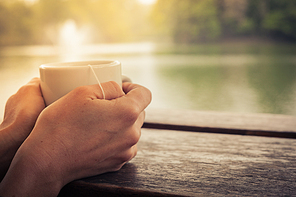 Closeup on a woman’s hands holding a cup of tea by a lake in the afternoon