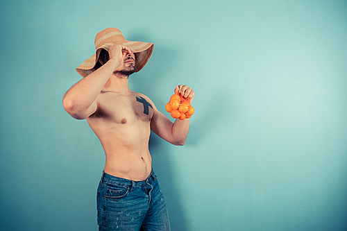 A young shirtless man wearing a large beach hat is holding a net of clementines