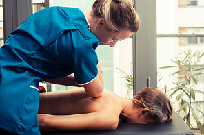 A massage therapist is treating a female client on a table by the window