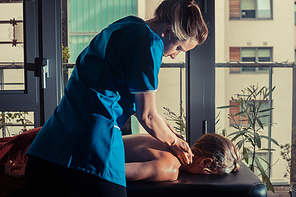 A massage therapist is treating a female client on a table by the window