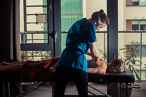 A massage therapist is treating a female client on a table by the window