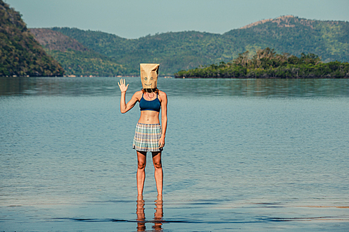 A silly young woman wearing a paper bag over her head is standing on a tropical beach and is waving