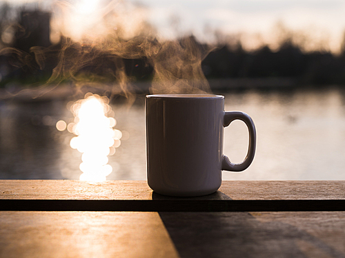 Steam rising from a mug with a hot beverage by a pond at sunset in the winter