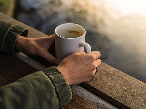 a young persons hands holding a mug with a hot beverage by a pond in a park at  in the winter
