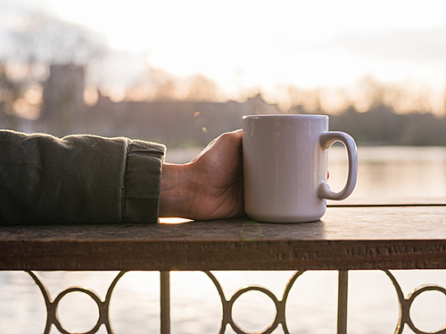 A young persons hands holding a mug with a hot beverage by a pond in a park at sunset in the winter