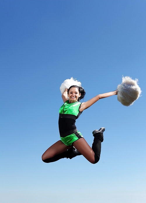 Young cheerleader in green costume jumping against blue sky