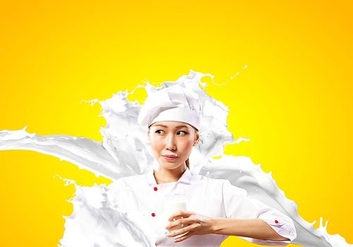 Asian female cook against milk splashes in red apron against color background holding glass of milk