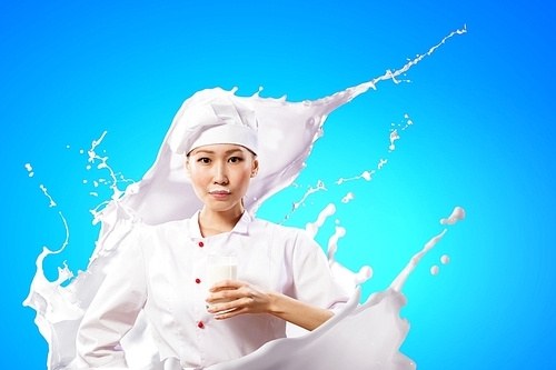 Asian female cook against milk splashes in red apron against color background holding glass of milk