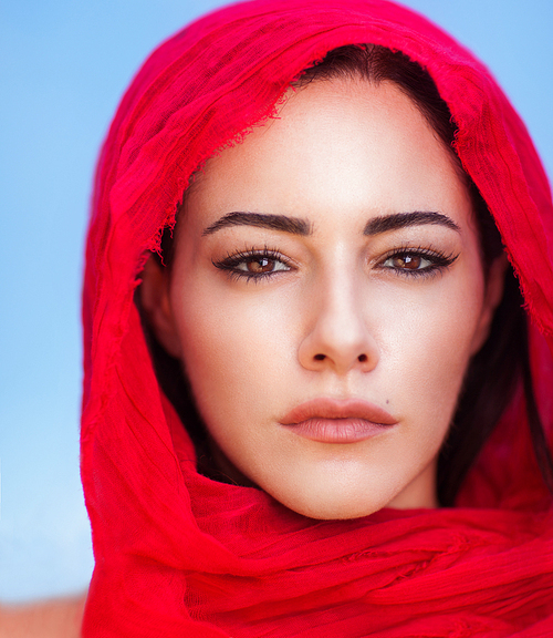 Closeup portrait of beautiful arabic woman wearing red headscarf over blue sky background, perfect natural makeup, traditional arabian beauty