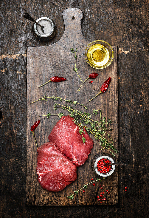 Raw meat steaks on wooden cutting board with oil, herbs and spices. Dark rustic background, top view