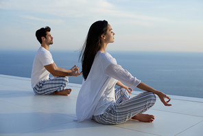 young couple practicing yoga at  in modern home terace with ocean and sunset in background